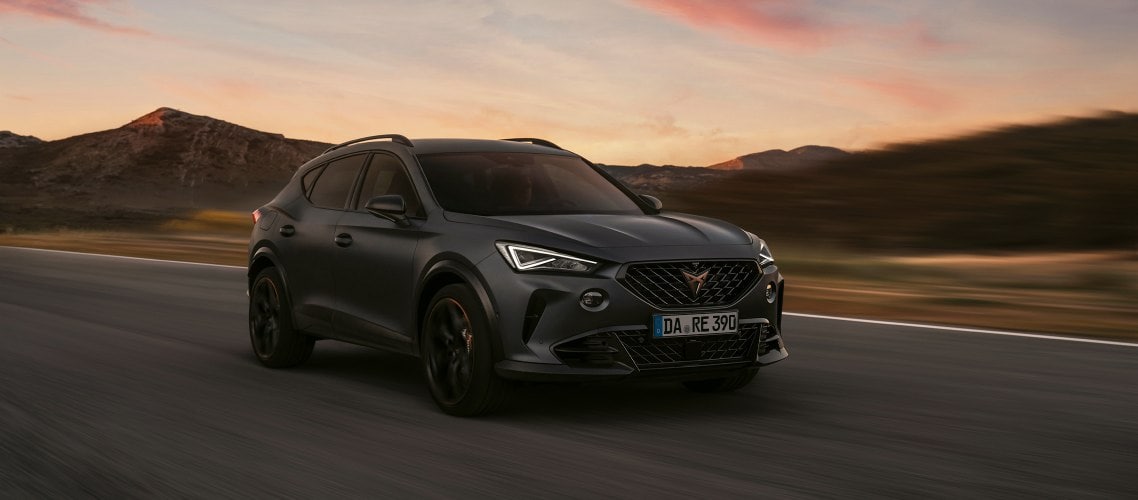 Der CUPRA Formentor  VZ von vorne, fahrend auf einer Straße. Im Hintergrund ein Sonnenuntergang mit Bergen.
