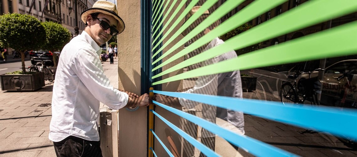CUPRA City Garage in München. Der Tape-Künstler Felix Rodewaldt  bringt gerade Regenbogenfarben Tapes auf den Fenstern an. Außenansicht.