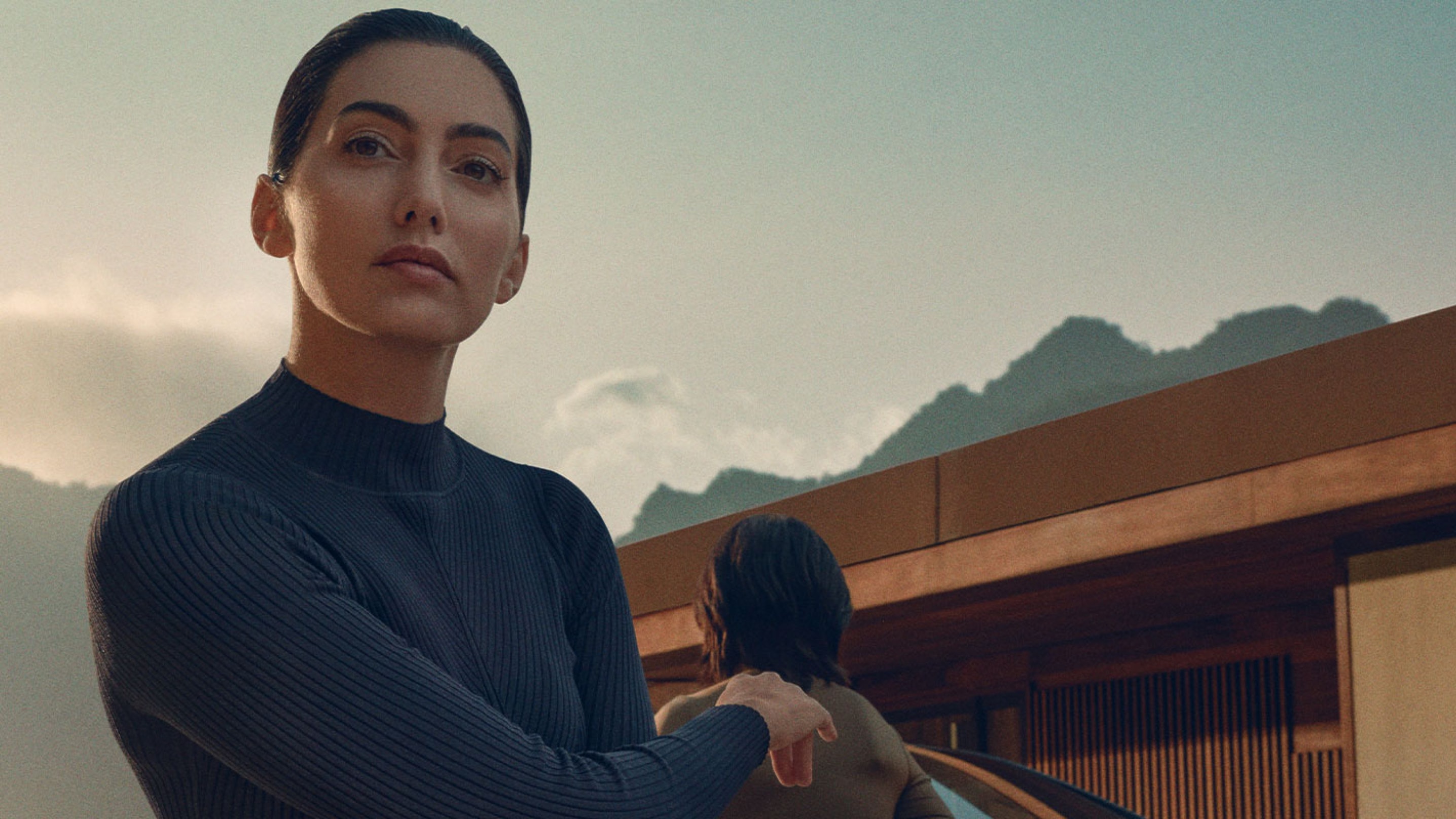 Eine Frau steht vor einem modernen Gebäude und schaut in die Kamera. Im Hintergrund ist der blaue Himmel und Berge zu erkennen.