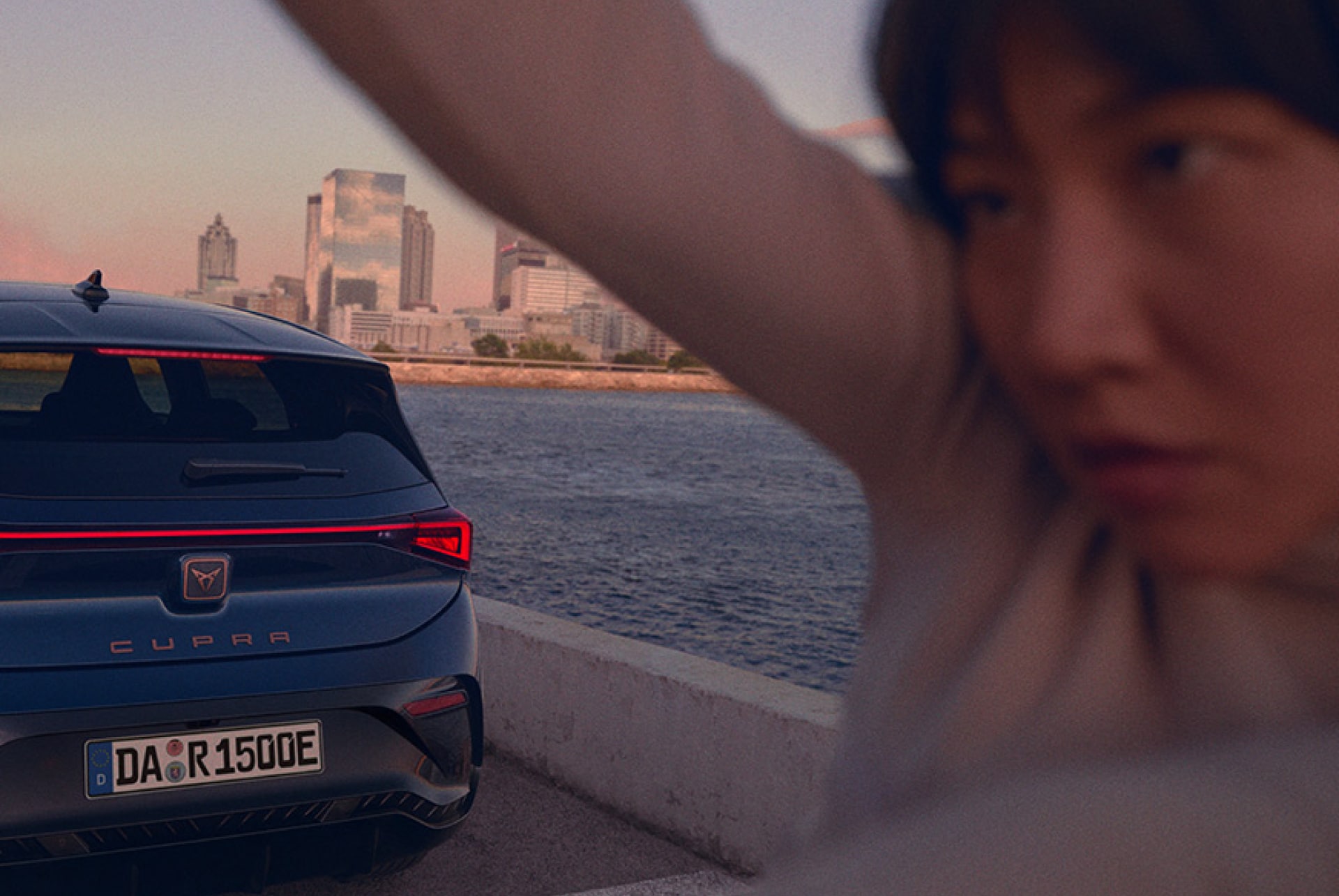 Ein blauer CUPRA. Born steht an einer Uferpromenande. Man erkennt die Hälfte der Rückseite des Autos und vor ihm eine Frau. Im Hintergrund ist das Meer, Strand und Hochhäuser zu erkennen.