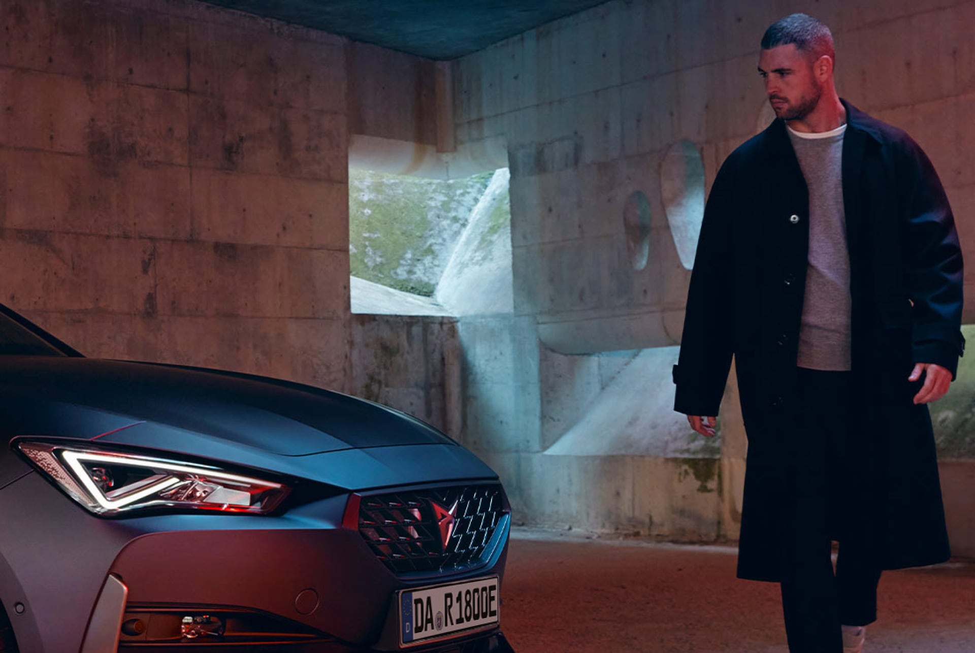 Ein Mann im minimalistischen Betonraum steht vor einem CUPRA Leon e-HYBRID – markante Lichtsignatur und progressives Design im Fokus.
