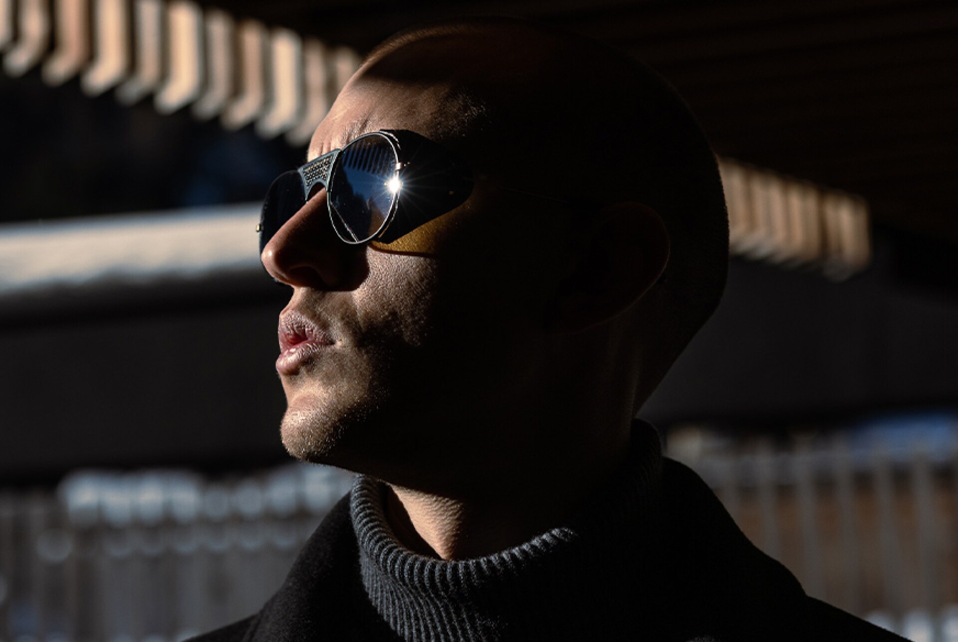 Seitenprofil eines Mannes mit Sonnenbrille, der in die Sonne sieht. Im Hintergrund ist ein modernen Gebäude zu erkennen.