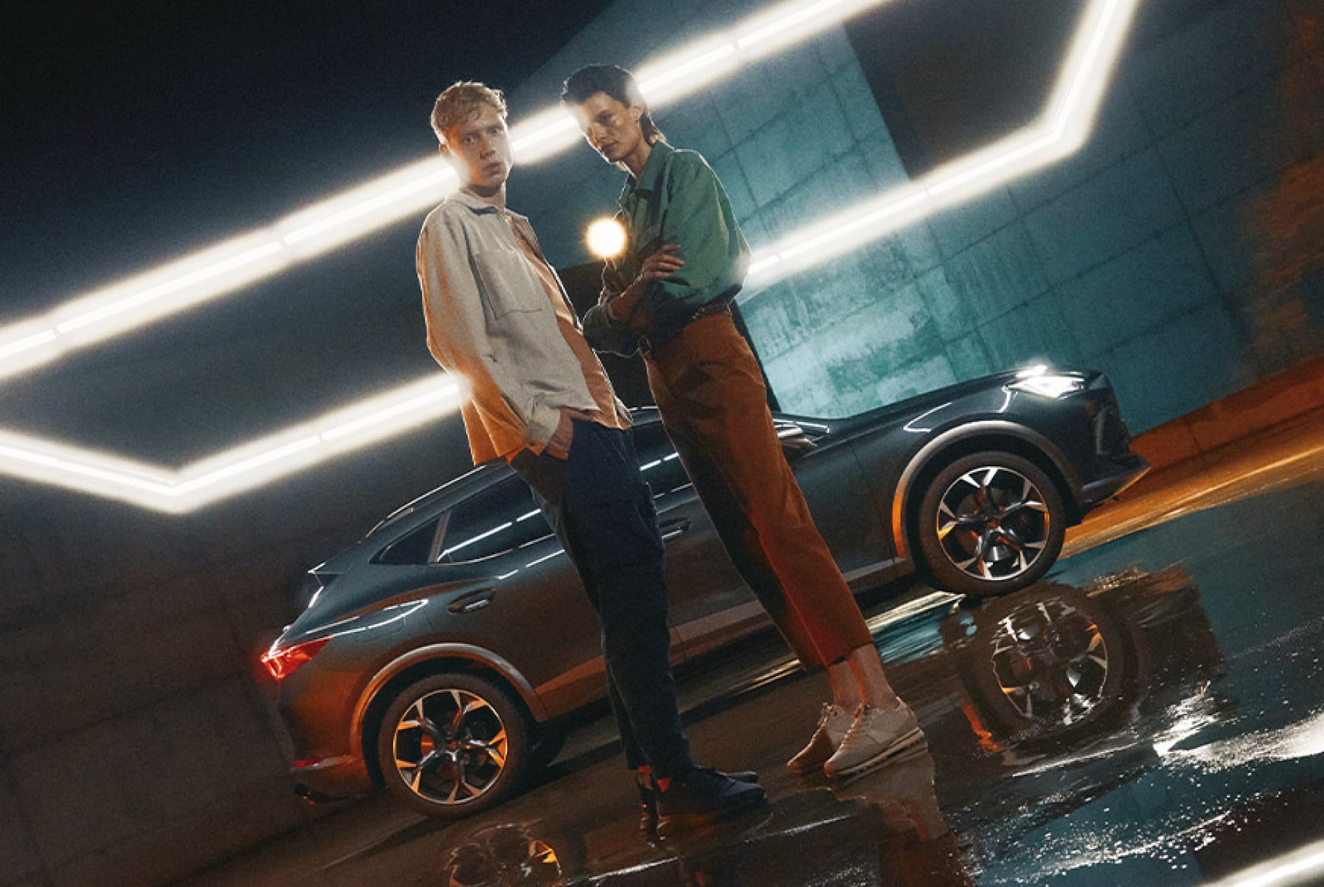 Seitenansicht eines CUPRA Fahrzeugs in Grau unter einer hellen Neon Röhre. Vor ihm stehen ein Mann und eine Frau und sehen in die Kamera.