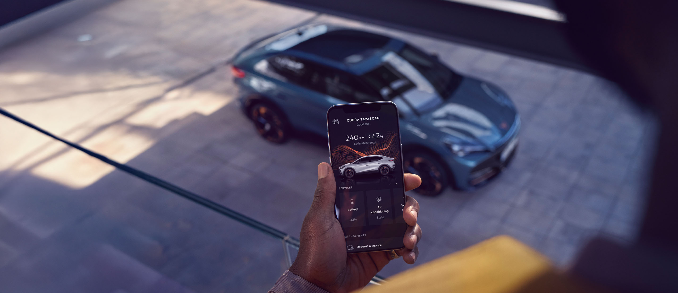 Ein CUPRA Tavascan steht auf einem Parkdeck. Im Vordergrund hält eine Person ihr Smartphone, mit der geöffneten My CUPRA App.  