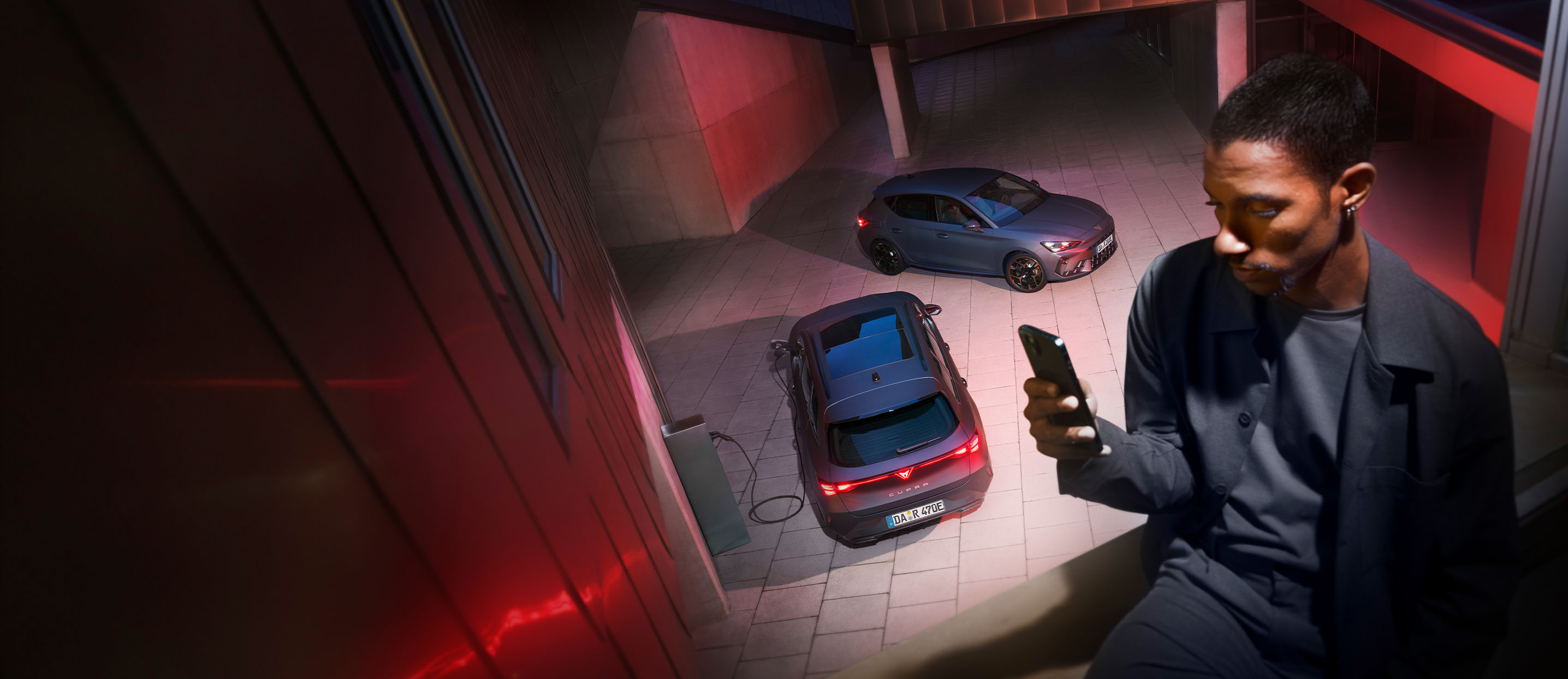 Mann sitzt am Fenster und steuert per Smartphone die Ladung eines CUPRA Elektrofahrzeugs, während zwei CUPRA Modelle in einem modernen Innenhof stehen.