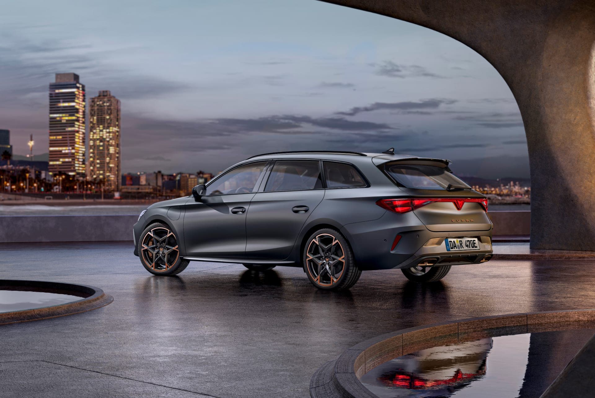 CUPRA Leon Sportstourer 2024 in Magnetic Grau, geparkt auf einer eleganten, dunklen Betonfläche unter einer modernen, geschwungenen Brücke. Im Hintergrund ist die beleuchtete Skyline von Barcelona bei Dämmerung zu sehen. Das Fahrzeug zeichnet sich durch sein sportliches Design und die auffälligen orangefarbenen Akzente an den Felgen aus. Die Rückleuchten und die dynamische Silhouette des Autos sind deutlich hervorgehoben.