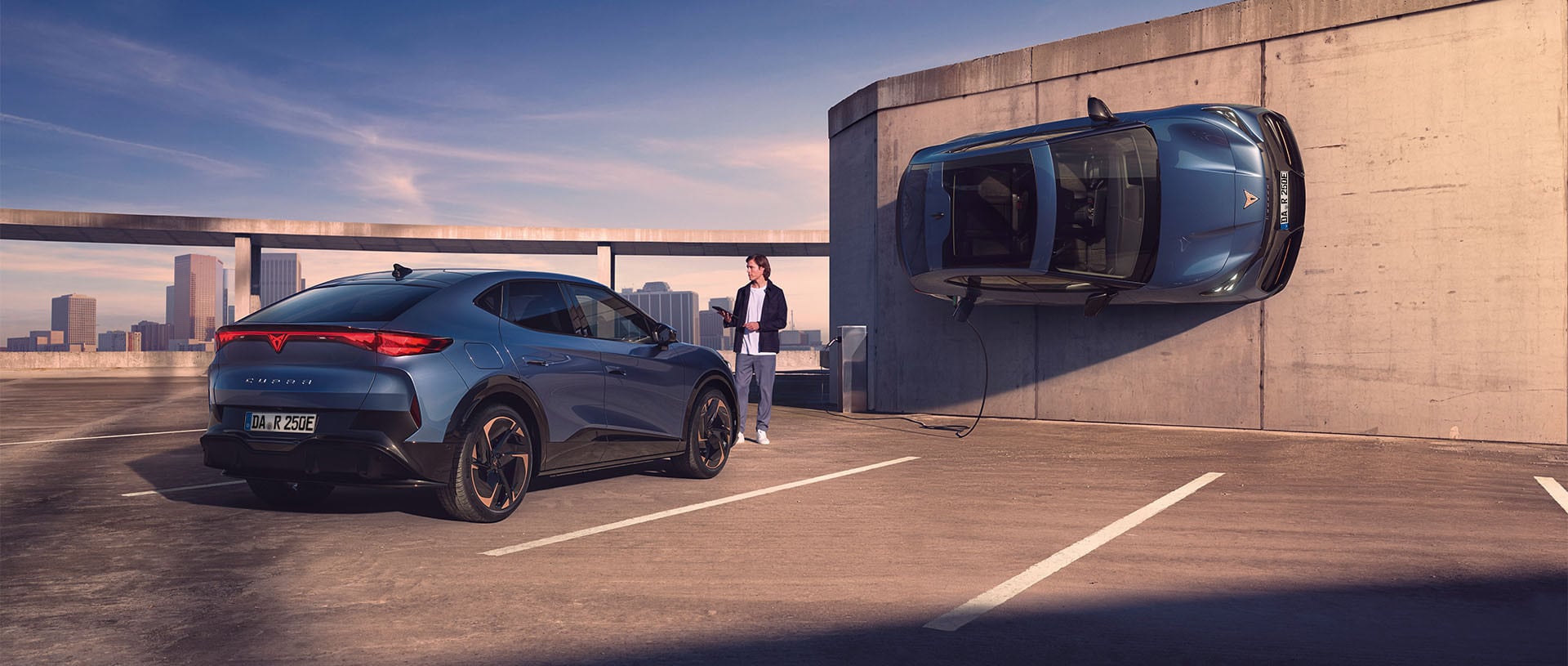 Zwei CUPRA Tavascan Elektrofahrzeuge stehen auf einem Parkdeck, eines wird über ein Ladekabel geladen, das andere ist vertikal an einer Betonwand positioniert.