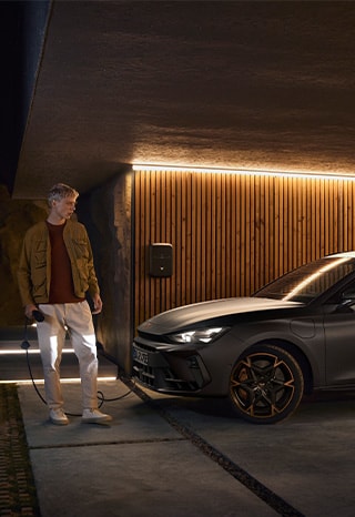 Ein%20CUPRA%20steht%20vor%20einer%20braunen%20Holzwand%20und%20wird%20mit%20einer%20Wallbox%20geladen.%20Vor%20den%20Scheinwerfern%20steht%20ein%20Mann%20mit%20dem%20Ladegerät%20in%20der%20Hand.