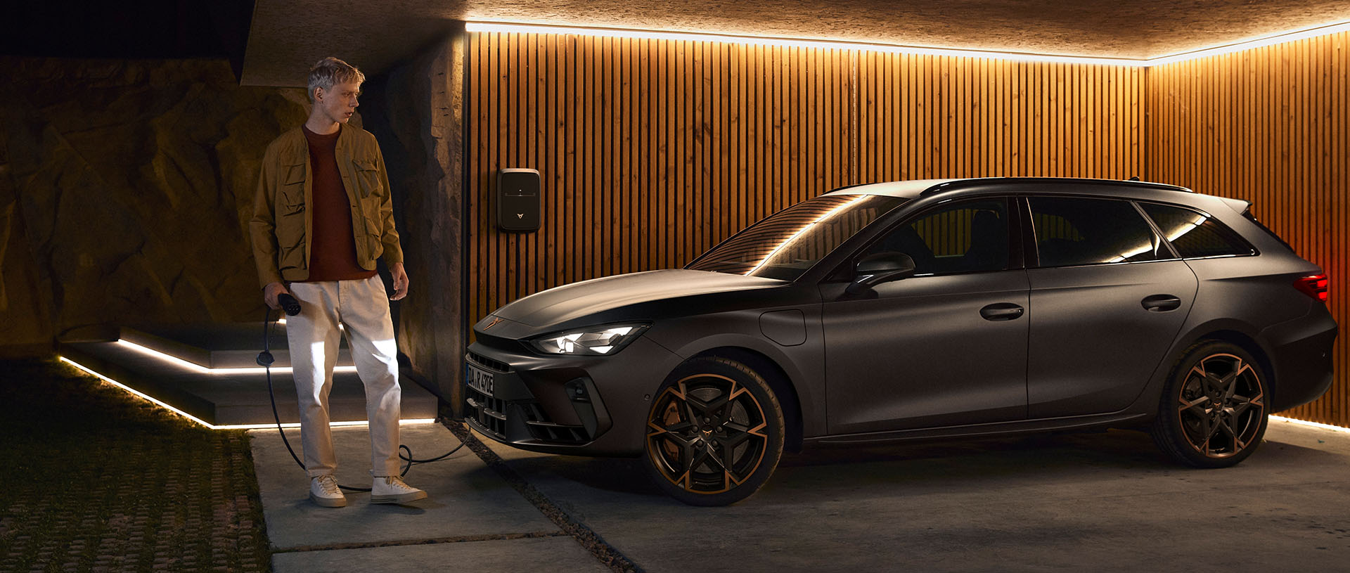 Ein CUPRA steht vor einer braunen Holzwand und wird mit einer Wallbox geladen. Vor den Scheinwerfern steht ein Mann mit dem Ladegerät in der Hand.