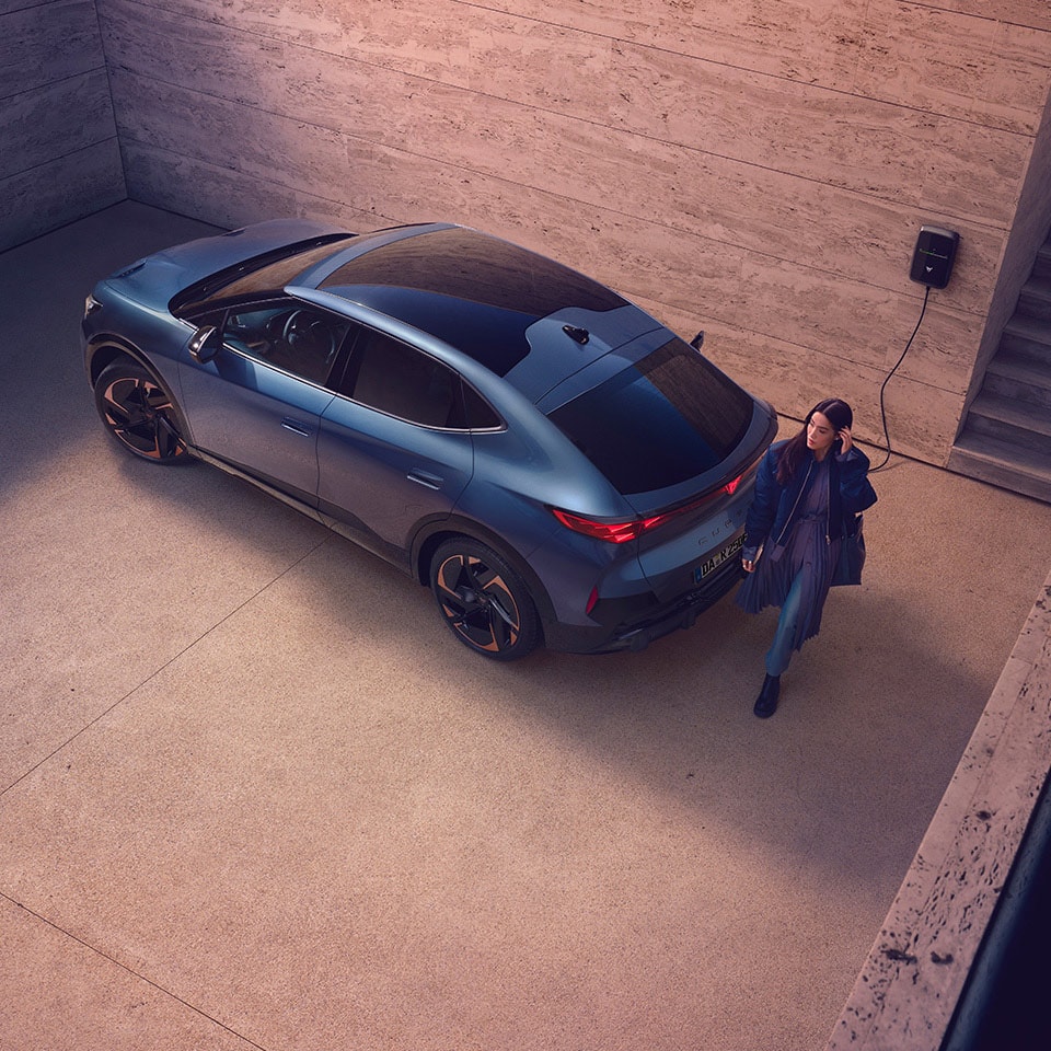 Ein CUPRA Tavascan steht vor einer beigen Wand und wird an einer Wallbox aufgeladen. Hinter dem CUPRA läuft eine Frau an der Heckklappe vorbei.