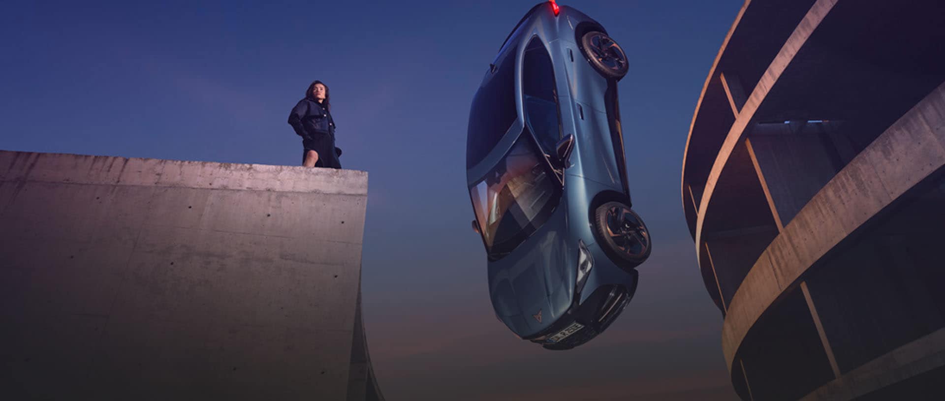 CUPRA Tavascan hängt an einem roten Faden vom Himmel herunter. Im Hintergrund sieht man eine Frau und ein rundes Gebäude.