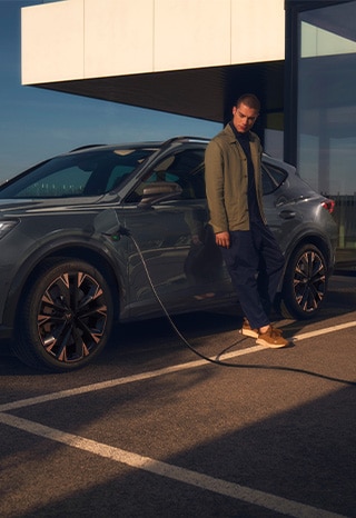 CUPRA%20Formentor%20ist%20an%20einer%20Ladesäule%20angeschlossen,%20urbaner%20Hintergrund%20mit%20Glas-%20und%20Betonelementen,%20Mann%20blickt%20an%20Fahrzeug%20lehnend%20auf%20die%20Ladeklappe