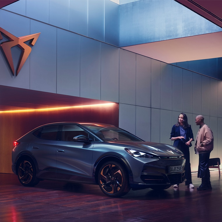 Eine stilvolle Szene mit dem CUPRA Tavascan in einer modernen Garage. Zwei Frauen stehen daneben und unterhalten sich, während das Fahrzeug an einer Ladestation angeschlossen ist. 