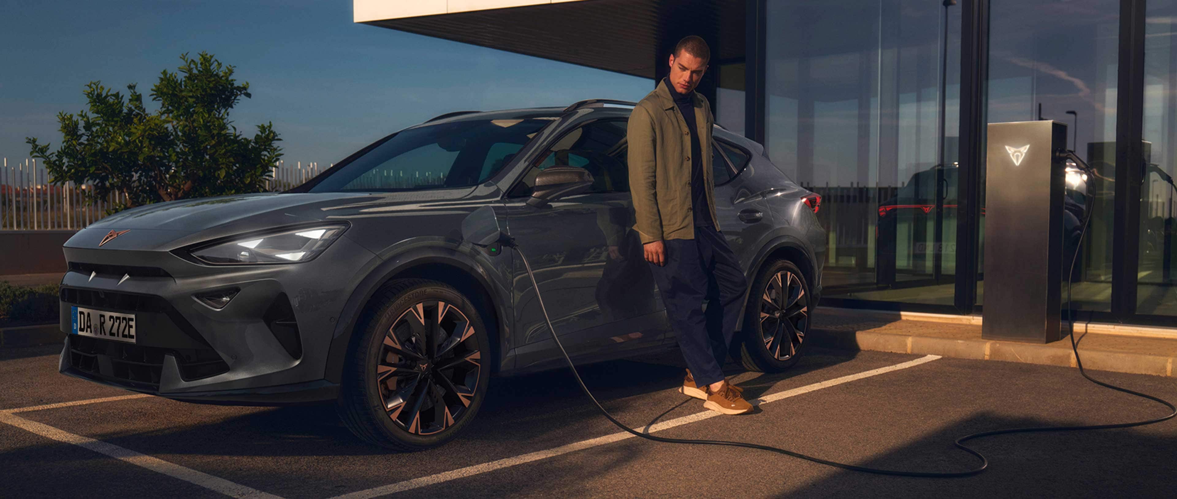 Ein Mann lehnt an einem CUPRA, während dieser an einer öffentlichen Ladestation auflädt. Im Hintergrund ist ein verglastes Gebäude zu erkennen.