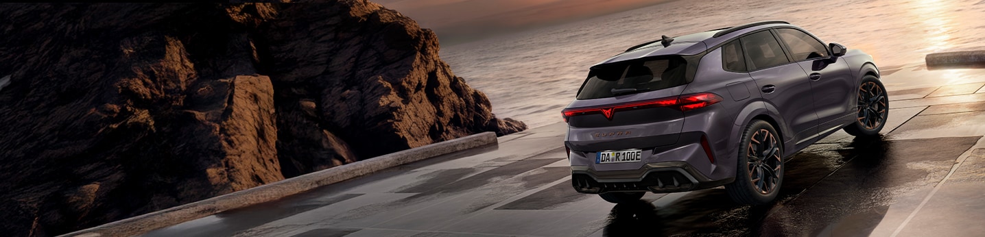 Ein CUPRA Leon und ein CUPRA Leon Sportstourer Plug-in-Hybrid in Mattgrau stehen auf einem beleuchteten Parkdeck mit E-Ladestation. Eine Verkäuferin zeigt einem Interessenten Details auf einem Tablet. Im Hintergrund erstreckt sich eine nächtliche Stadtlandschaft.