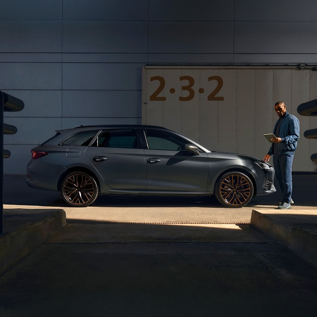 Ein CUPRA Leon Sportstourer in mattem Grau steht seitlich vor einem modernen Industriegebäude. Rechts neben dem Fahrzeug steht ein Mann in dunkler Kleidung, der auf ein Tablet schaut.