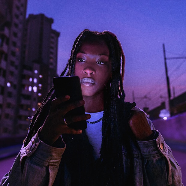 Eine junge Frau mit langen Dreadlocks steht nachts in einer urbanen Umgebung und blickt konzentriert auf ihr Smartphone. Im Hintergrund sind Hochhäuser mit leuchtenden Fenstern und eine Bahnlinie bei violett-blauem Himmel zu sehen.