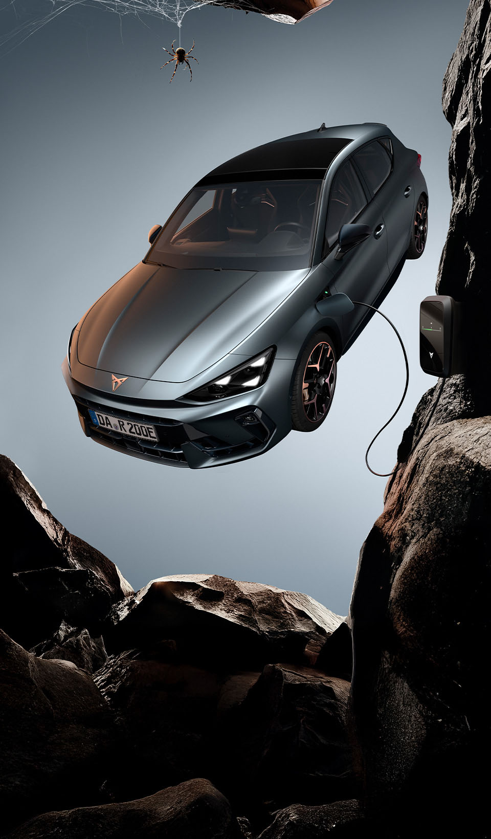 Ein CUPRA Leon Hatchback, von oben gesehen, mit eingeschalteten Scheinwerfern, neben einer schwarzen Spinne, die von einem Felsvorsprung herunterhängt. Ein Ladekabel ist von einer Ladeeinheit an der Klippe mit dem Auto verbunden.