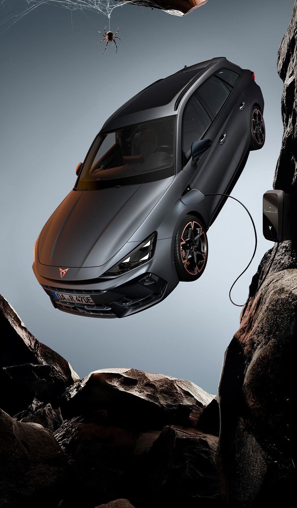 Ein CUPRA Leon Sportstourer, von oben gesehen, mit eingeschalteten Scheinwerfern, neben einer schwarzen Spinne, die von einem Felsvorsprung herunterhängt. Ein Ladekabel ist von einer Ladeeinheit an der Klippe mit dem Auto verbunden.