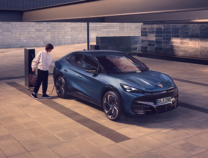 Eine Person lädt einen CUPRA Tavascan, ein vollelektrisches Auto, an einer öffentlich zugänglichen E-Ladestation in einer modernen urbanen Umgebung auf. Das sportliche Elektroauto steht auf einem hell gepflasterten Platz, im Hintergrund moderne Gebäude