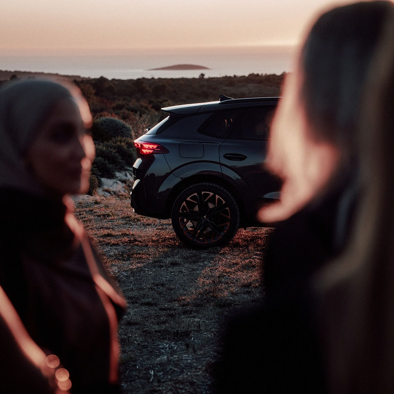 CUPRA Terramar steht in einer Landschaft bei Sonnenuntergang, im Vordergrund sind zwei Frauen unscharf zu sehen.