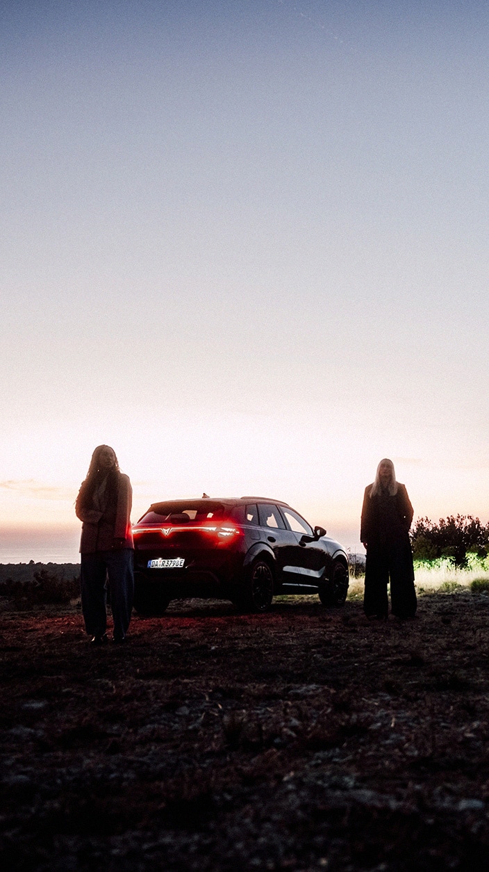CUPRA Terramar in natürlicher Umgebung, mit Gegenlicht inszeniert, Heckbeleuchtung strahlt rot. Models stehen Richtung Kamera