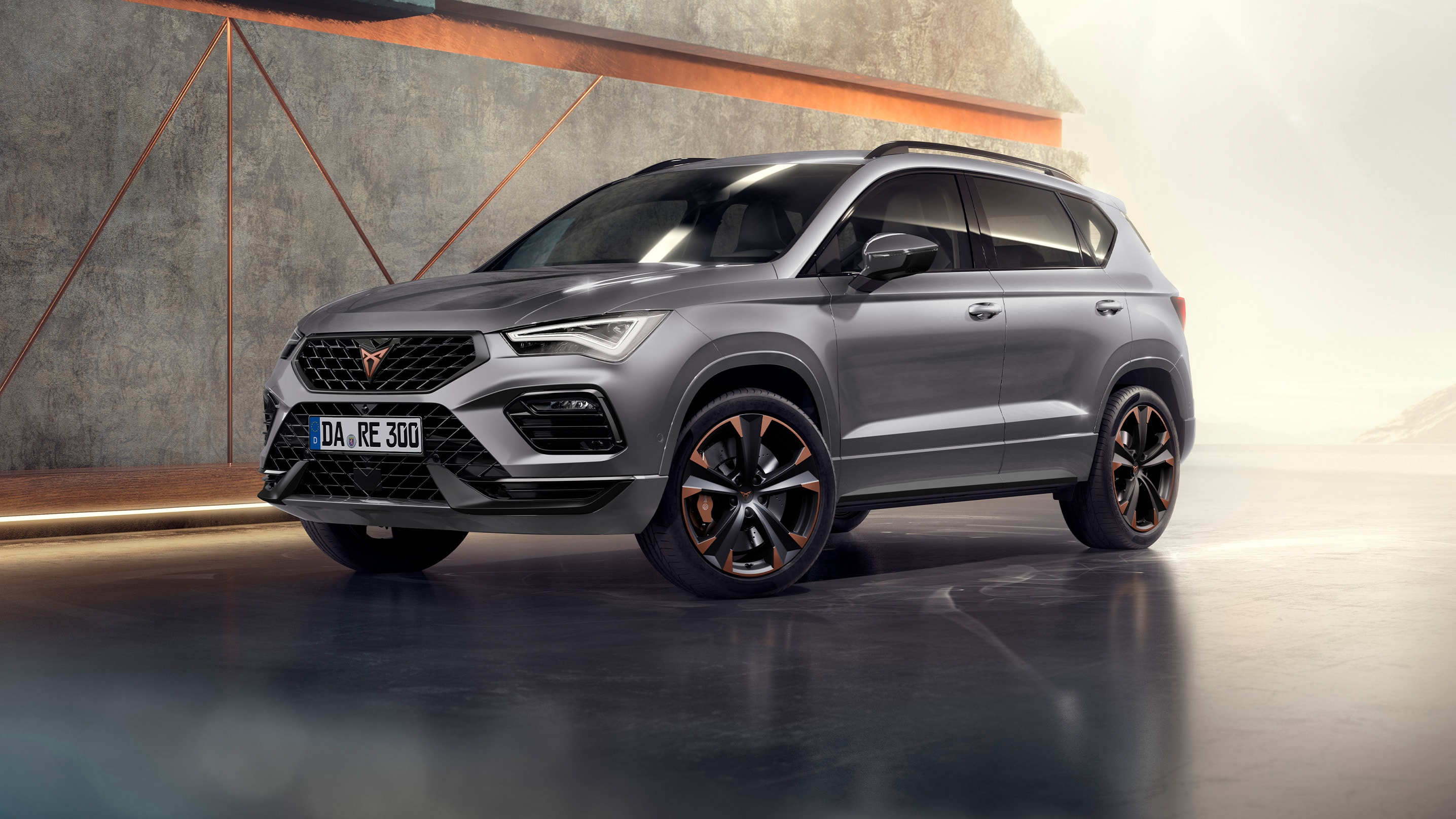 Ein CUPRA Ateca 2024 in Graphitgrau, ein sportliches und kompaktes SUV, geparkt in einer modernen, industriellen Umgebung. Das Fahrzeugdesign zeichnet sich durch aerodynamische Linien, LED-Scheinwerfer und markante Leichtmetallräder mit kupferfarbenen Akzenten aus. Der Hintergrund umfasst ein geometrisches Wanddesign, das einen Kontrast zum eleganten Fahrzeugdesign bildet.