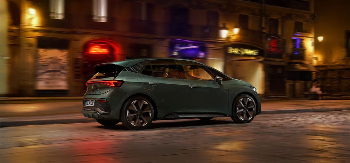 Ein grüner CUPRA Born VZ fährt dynamisch durch eine urbane Landschaft bei Sonnenuntergang.