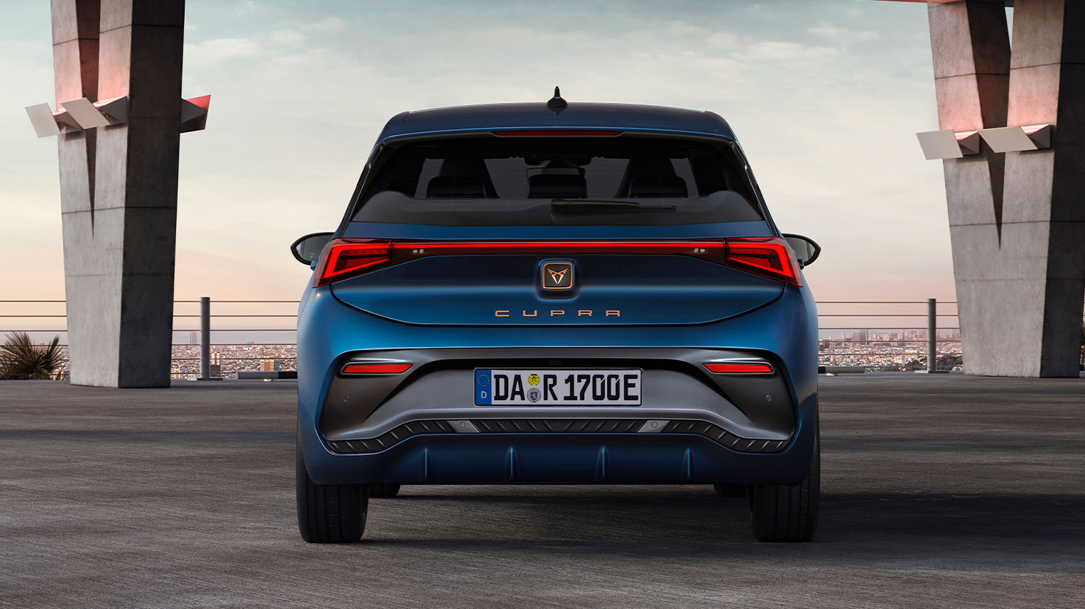 Der CUPRA Born in Aurora Blue aus der Heckperspektive, geparkt unter einer modernen Betonarchitektur mit Blick auf den Sonnenuntergang.