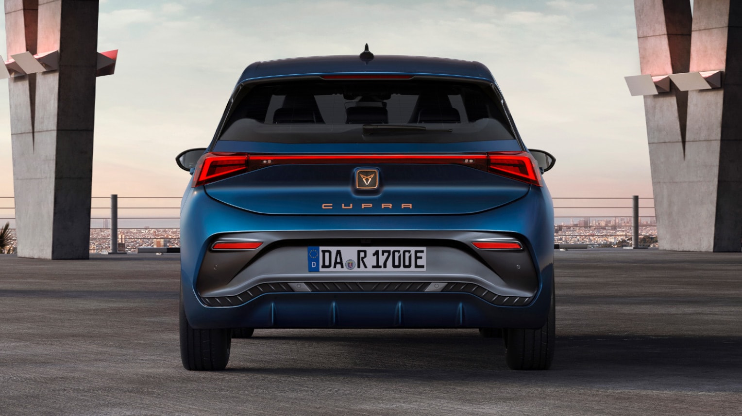 Der CUPRA Born in Aurora Blue aus der Heckperspektive, geparkt unter einer modernen Betonarchitektur mit Blick auf den Sonnenuntergang.