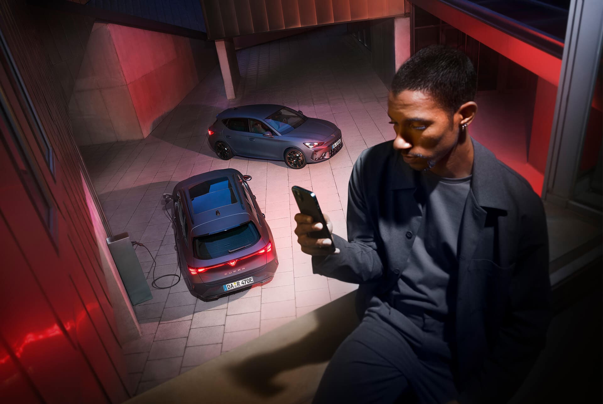 Ein Mann blickt auf sein Smartphone, während zwei CUPRA Modelle auf einem modernen, urbanen Platz stehen.