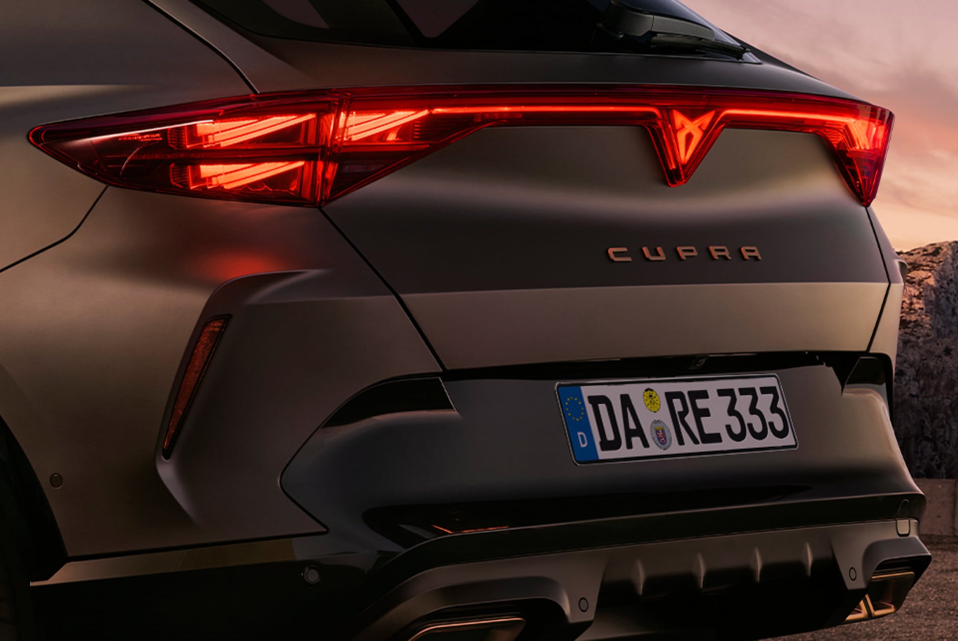 Hintere Dreiviertelansicht des CUPRA Formentor Hybrid, geparkt an der Uferpromenade vor der Skyline der Stadt. Das Fahrzeug ist so positioniert, dass das hintere Design, die Leuchten und die sportlichen Doppelauspuffrohre zur Geltung kommen.