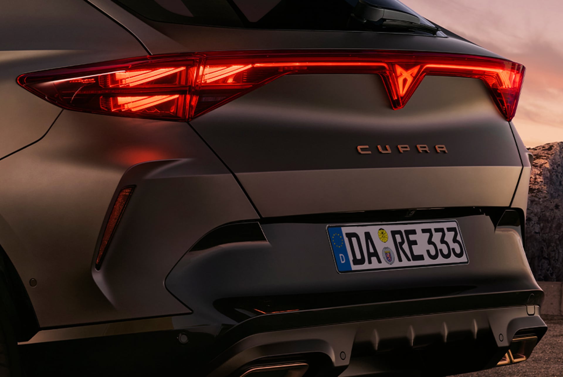 Hintere Dreiviertelansicht des neuen CUPRA Formentor 2024 Hybrid, geparkt an der Uferpromenade vor der Skyline der Stadt. Das Fahrzeug ist so positioniert, dass das hintere Design, die Leuchten und die sportlichen Doppelauspuffrohre zur Geltung kommen.