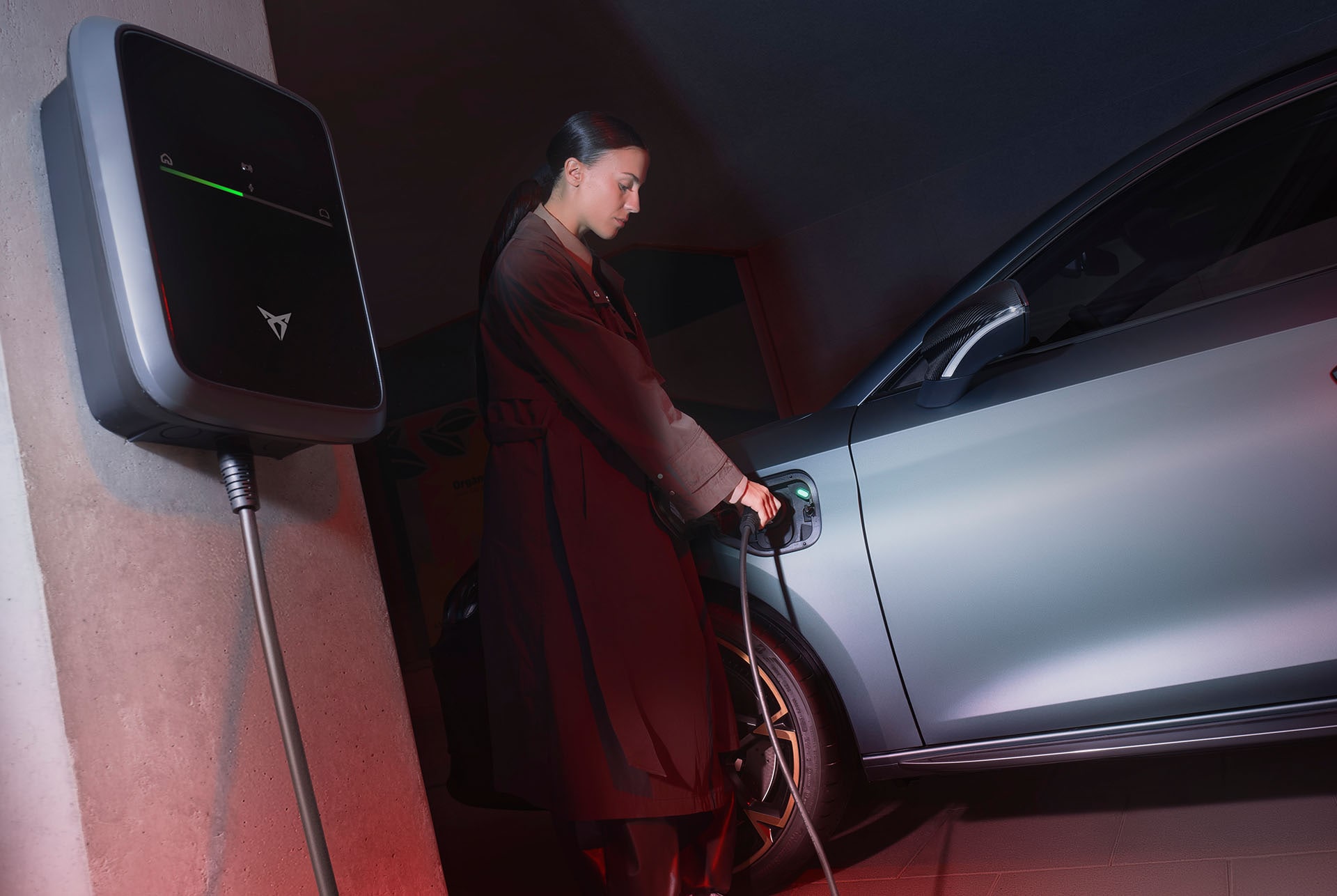 Eine Frau lädt einen CUPRA an einer Wallbox auf, während eine atmosphärische Beleuchtung die Szene akzentuiert.