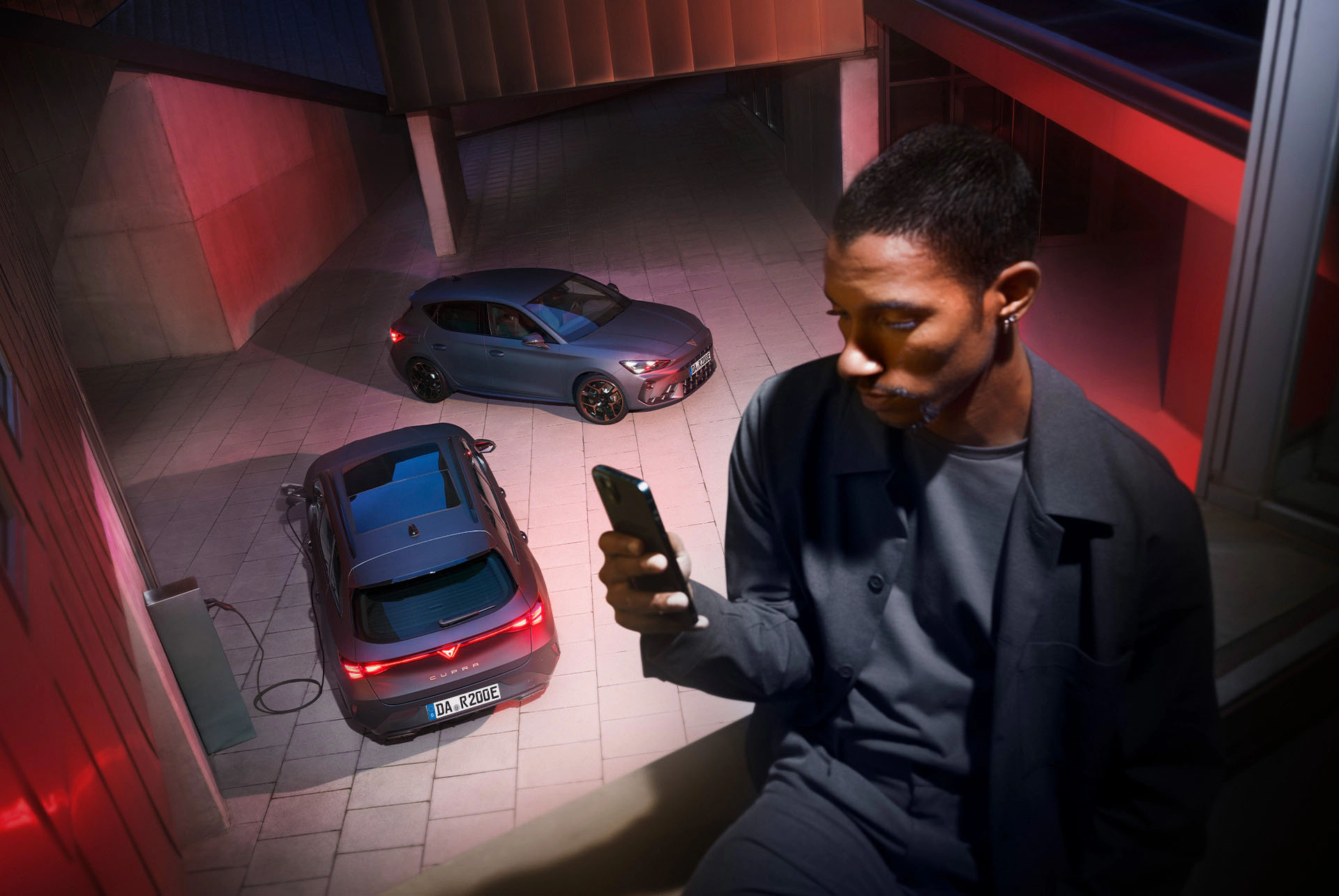 Ein Mann blickt auf sein Smartphone, während zwei CUPRA Modelle auf einem modernen, urbanen Platz stehen.