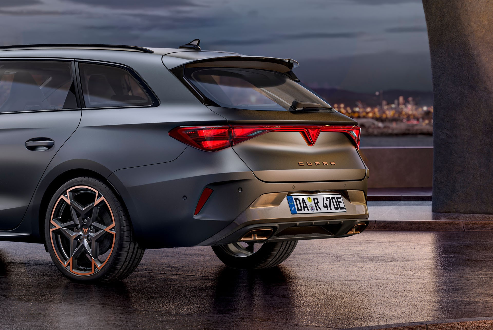 Close-up der Rückansicht des CUPRA Leon Sportstourer in Enceladus Grey Matt, mit Kupfer-Akzent und geschmiedeten Rädern, vor einer Betonstruktur und Skyline.