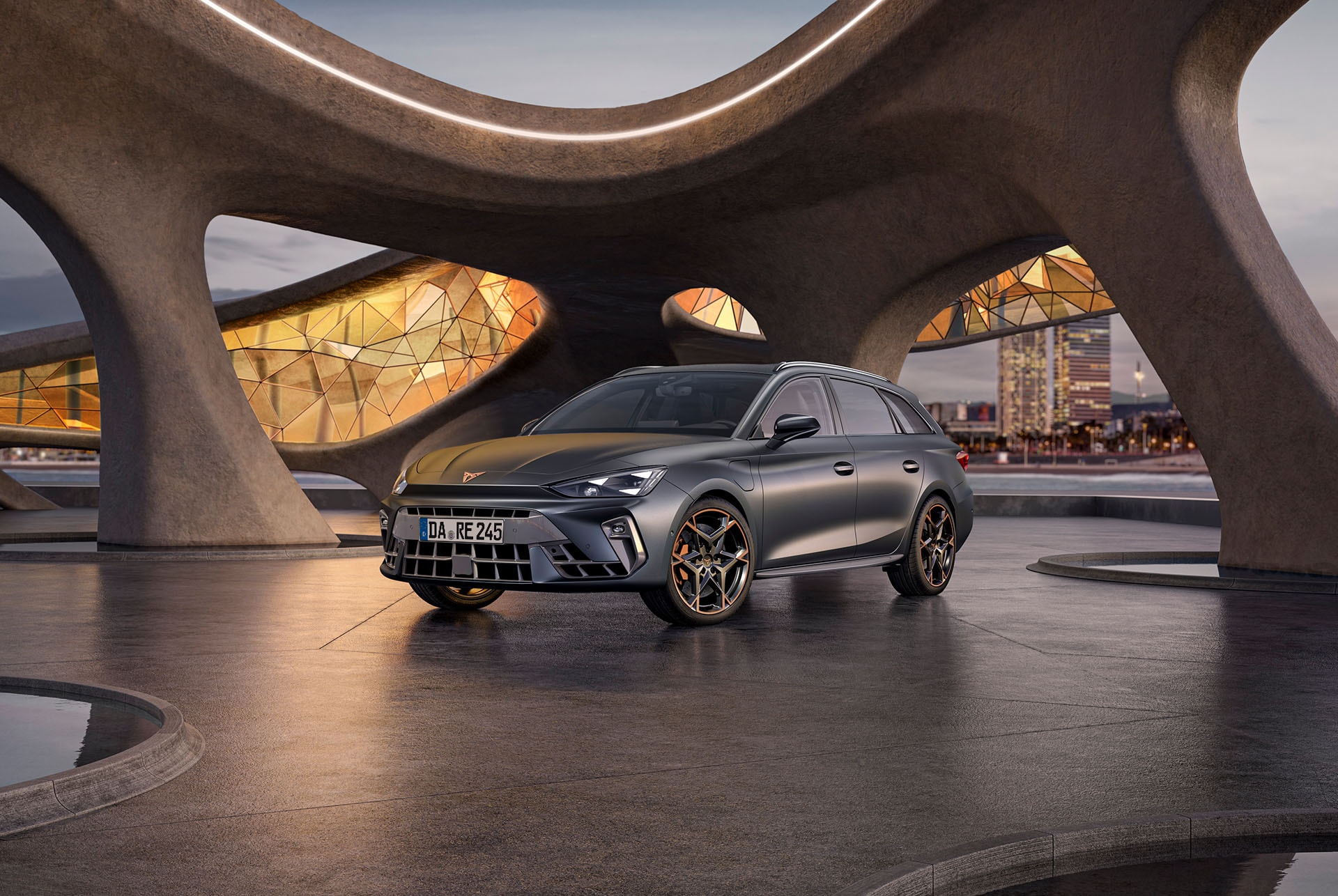 Seitenansicht des CUPRA Leon Sportstourer in Magnetic Grau, geparkt unter einer modernen, geschwungenen Betonstruktur. Das Fahrzeug sticht mit seinen auffälligen orangefarbenen Akzenten an den Felgen hervor. Im Hintergrund sind die beleuchtete Skyline und architektonische Highlights von Barcelona bei Dämmerung zu erkennen. 