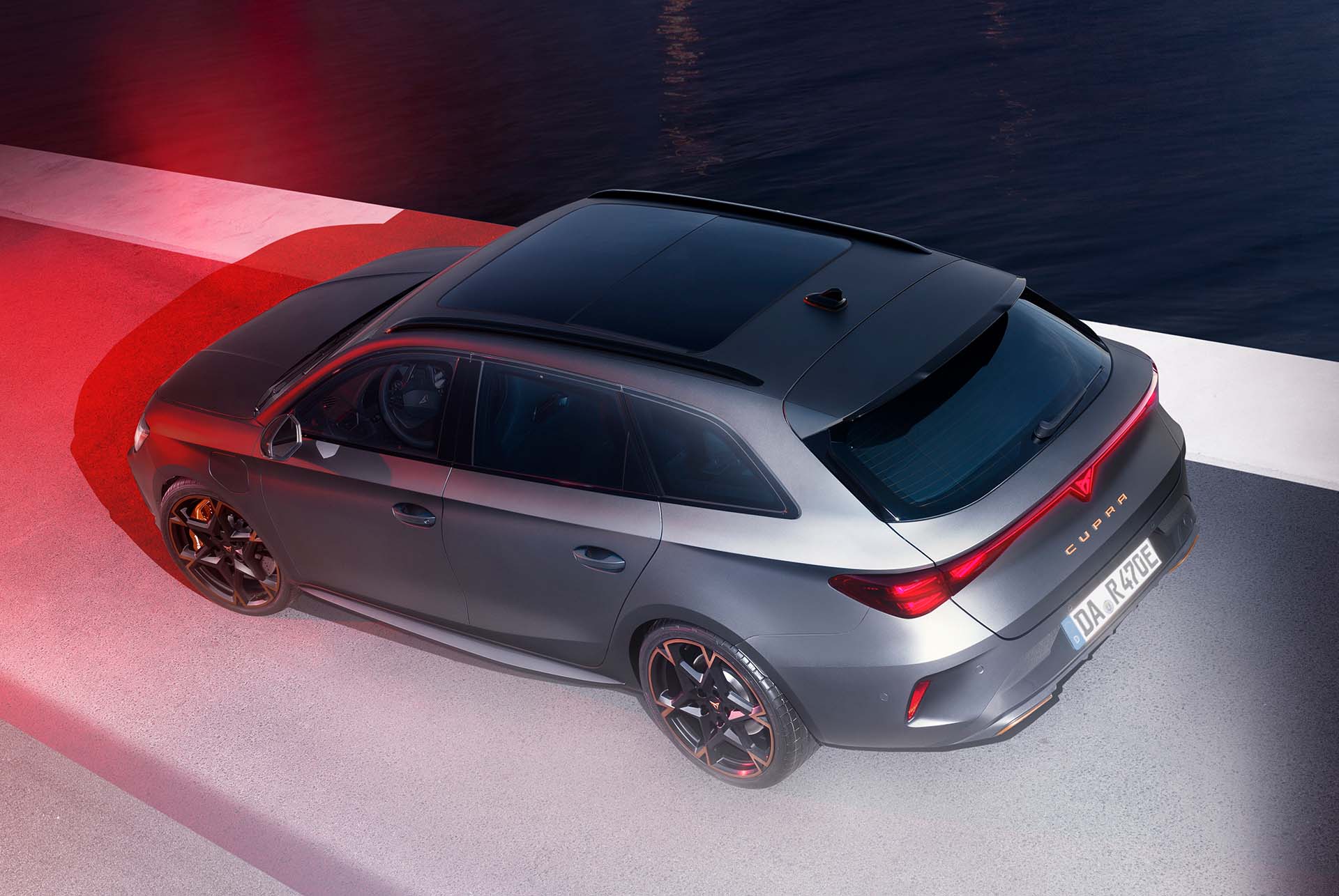 Das Bild zeigt einen CUPRA Leon Sportstourer bei Nacht an einer Küstenstraße mit Blick auf eine beleuchtete Stadt im Hintergrund. Das Fahrzeug ist in einer dunklen Metallic-Lackierung gehalten und wird durch rote und weiße Lichteffekte dramatisch in Szene gesetzt. Die moderne, sportliche Linienführung und das durchgehende LED-Rücklicht betonen die dynamische Erscheinung des Autos.