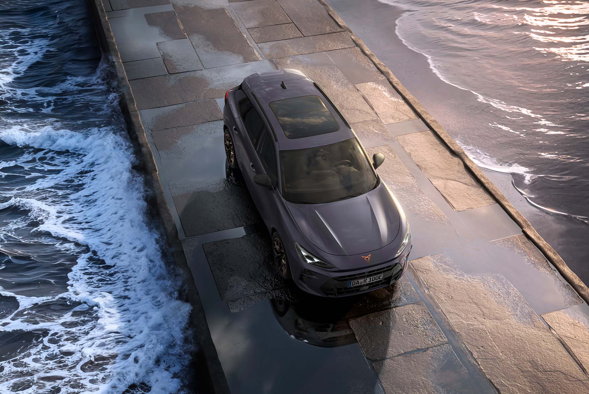 Der Cupra Terramar 2024 Hybrid Geländewagen mit Panoramadach, schlankem Design und modernen Leichtmetallrädern auf einem nassen Bürgersteig am Meer - aus der Vogelperspektive.
