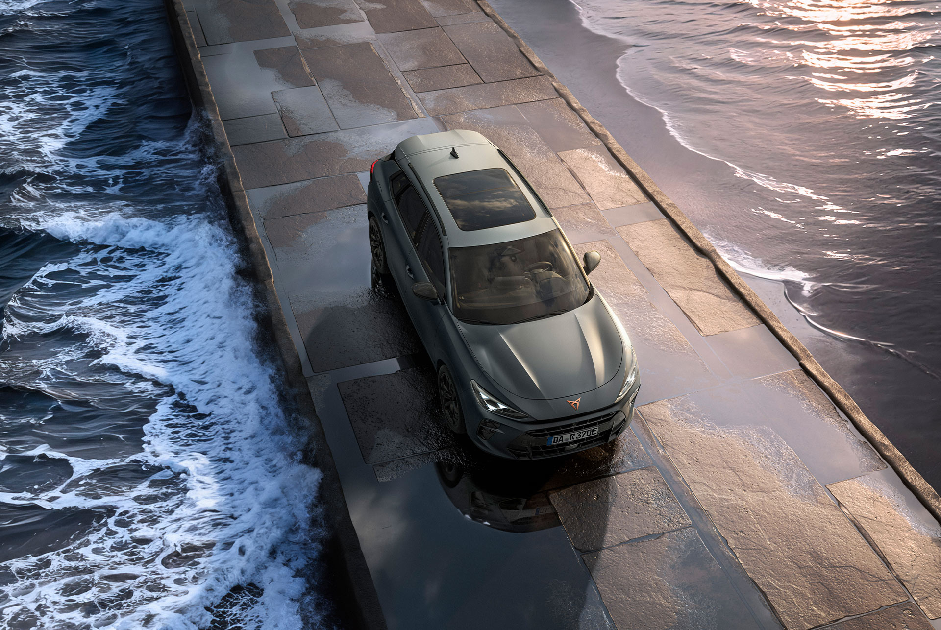 Der Cupra Terramar 2024 Hybrid Geländewagen mit Panoramadach, schlankem Design und modernen Leichtmetallrädern auf einem nassen Bürgersteig am Meer - aus der Vogelperspektive.