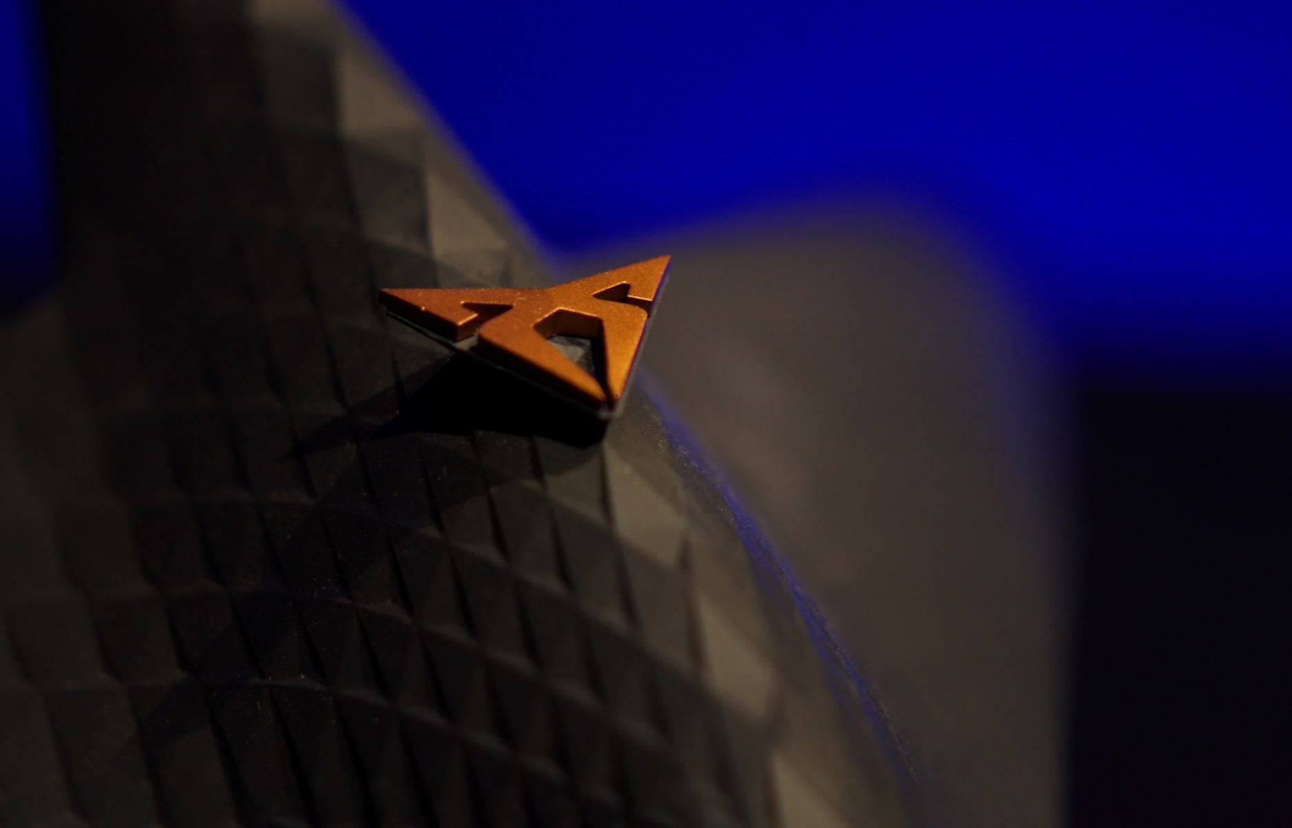 Kupfer farbenes CUPRA Logo vor einem schwarz-blauem Hintergrund.