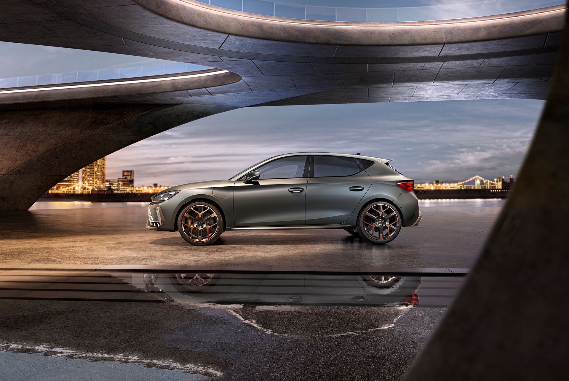 Weitwinkelaufnahme der Seitenansicht des grauen CUPRA Leon 2024 mit Mild-Hybrid-Antrieb, geparkt auf glattem Beton unter einer architektonischen Struktur mit Blick auf das Meer und die Skyline der Stadt, bei bewölktem Himmel.