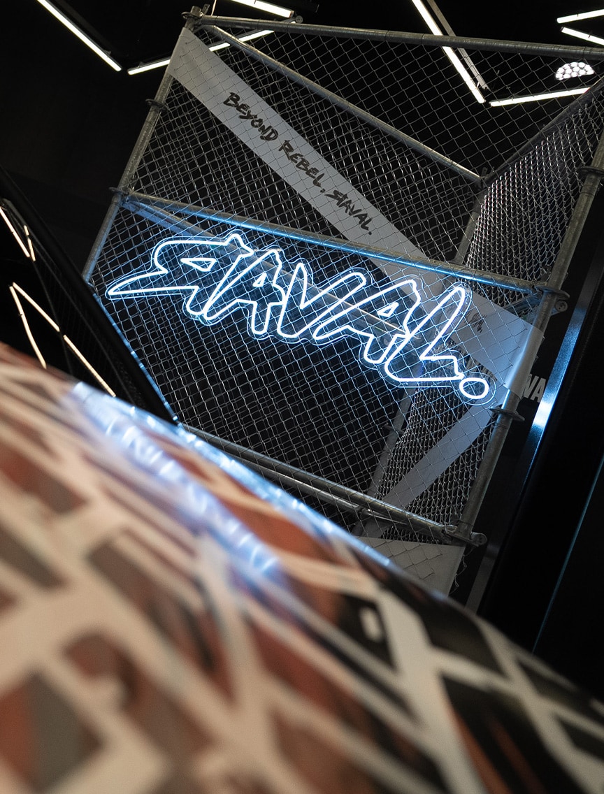 Auf einem Maschendrahtzaun ist eine Leuchtreklame mit der Aufschrift „RAVAL!“ angebracht, auf der auch der Text „BEYOND REBEL, RAVAL.“ zu lesen ist. Die Anzeige befindet sich in einem dunklen, stilisierten Raum mit geometrischen Lichtmustern.