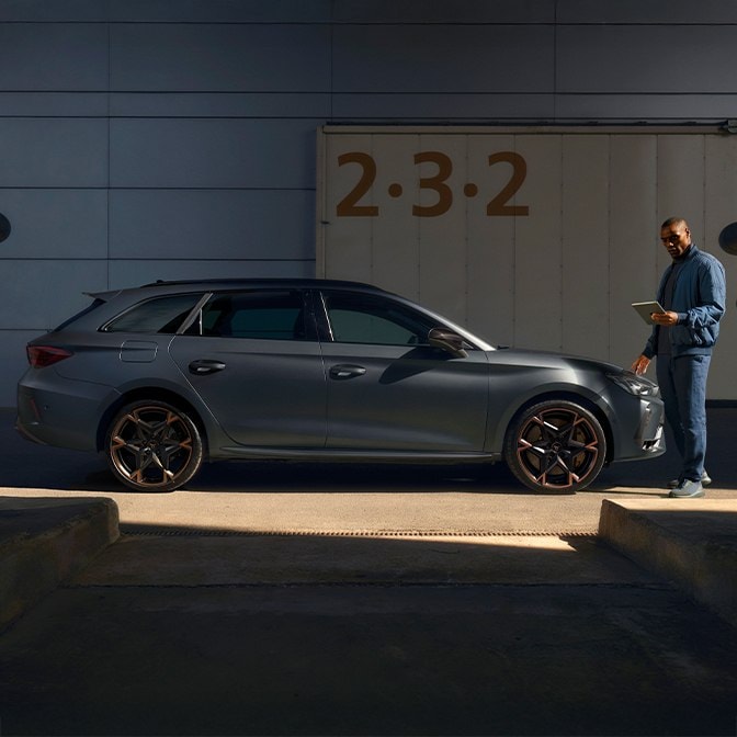 Eine Seitenansicht eines mattgrauen CUPRA Leon mit den charakteristischen Leichtmetallfelgen mit Kupferakzenten, der in einer betonierten Industrie-Laderampe geparkt ist. Ein Mann in einer blauen Jacke steht vor dem Fahrzeug und blickt auf ein Tablet, während das Sonnenlicht scharfe Schatten über die Szene wirft.