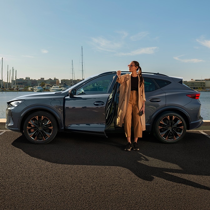 Eine Frau lehnt an einem grauen CUPRA Leon und schaut auf ihr Handy, während das Auto vor einem modernen, minimalistischen Haus aus Holz und Beton an einer Wandladestation angeschlossen ist.