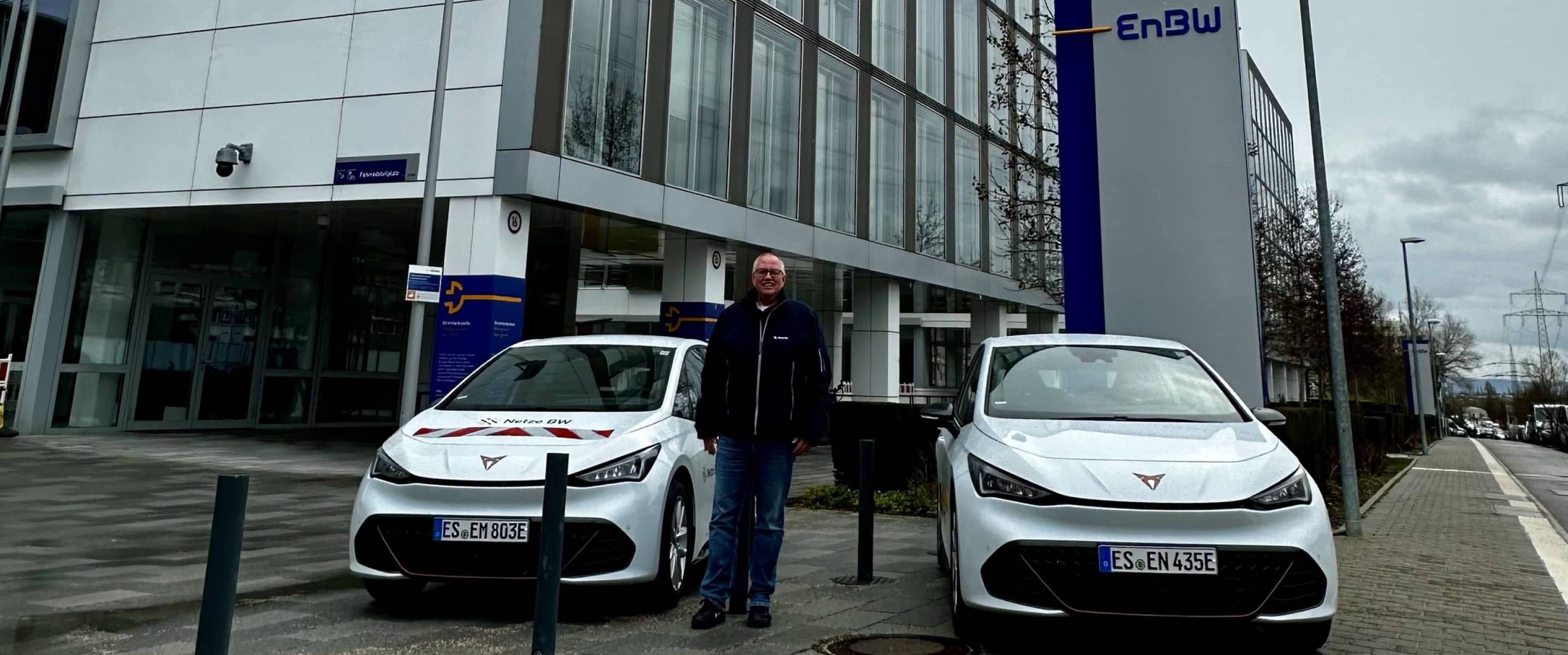 Zwei weiße CUPRA-Fahrzeuge, die vor einem enbw-Bürogebäude geparkt sind, mit einem Fuhrparkmanager, der an einem bewölkten Tag zwischen den Autos steht.