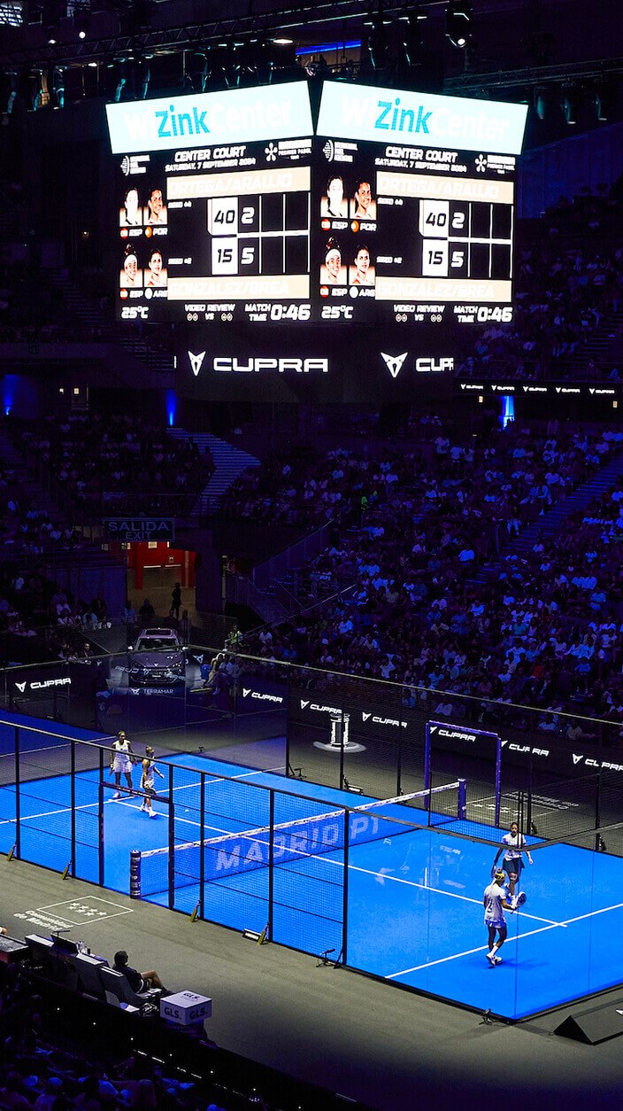 Ein Padel-Platz, beleuchtet von blauen Lichtern, von oben gesehen während eines Spiels in einem großen Stadion voller Zuschauer. Zwei Spielerpaare befinden sich auf dem Platz, und im Hintergrund ist eine große Anzeigetafel zu sehen.