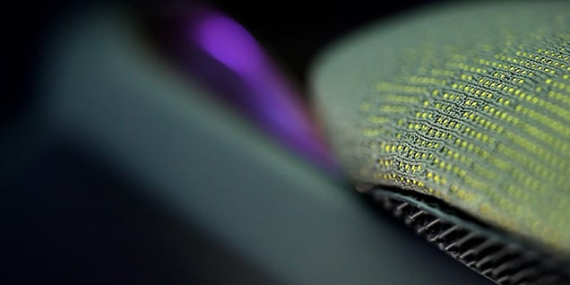 Das Bild zeigt eine Makroaufnahme einer strukturierten Oberfläche. Der Fokus liegt auf einem Material mit einem gewebten oder gestrickten Muster, bei dem kleine gelbe Fäden mit grauen oder grünlichen Fasern verflochten sind.
