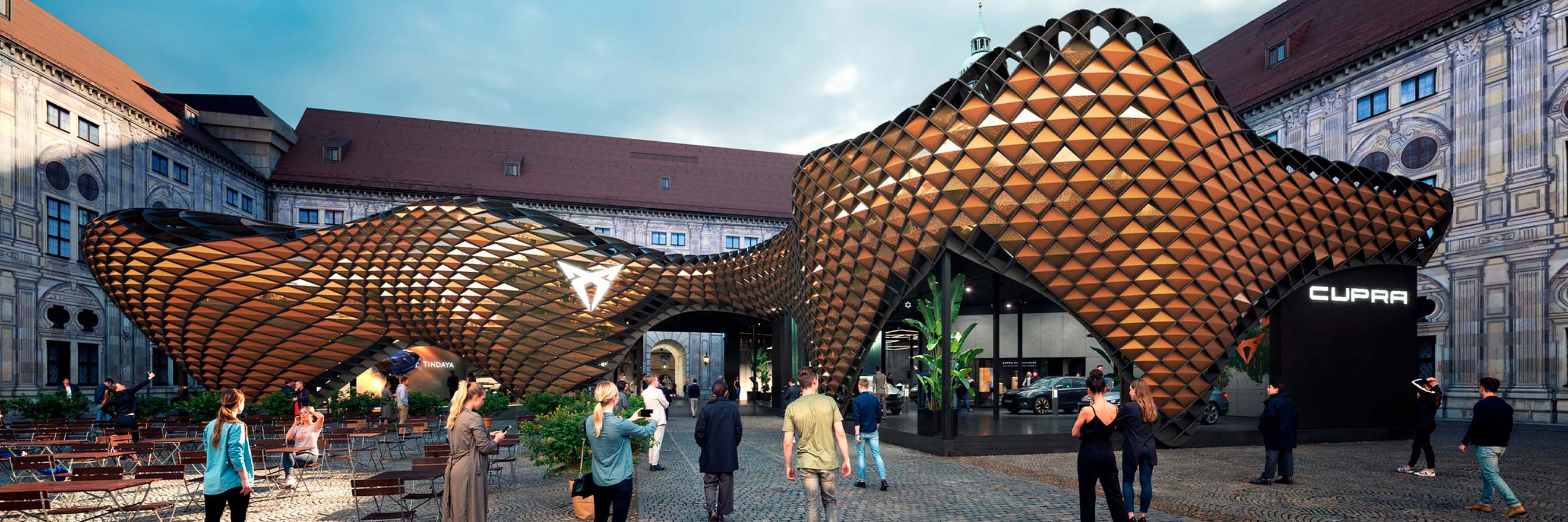 Eine moderne, wellenförmige Architekturstruktur mit einem Muster aus miteinander verbundenen, metallischen Diamantformen dominiert die Bildmitte. Die Struktur ist ein Pavillon im Freien mit einer großen Öffnung in der Mitte, die ein Cupra-Logo und einen Blick auf einen Innenraum freigibt.