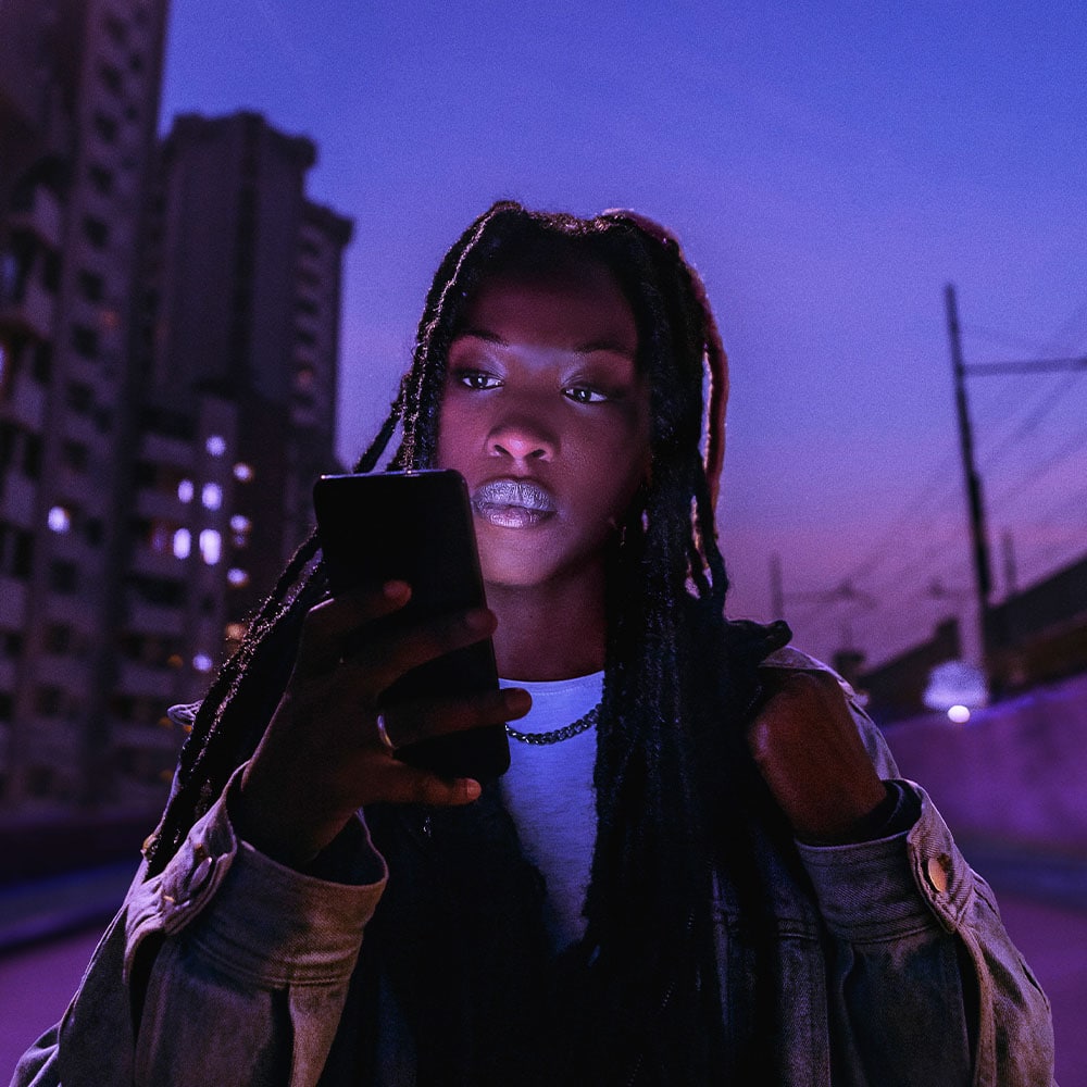 Eine junge Frau mit langen Dreadlocks steht nachts in einer urbanen Umgebung und blickt konzentriert auf ihr Smartphone. Im Hintergrund sind Hochhäuser mit leuchtenden Fenstern und eine Bahnlinie bei violett-blauem Himmel zu sehen.